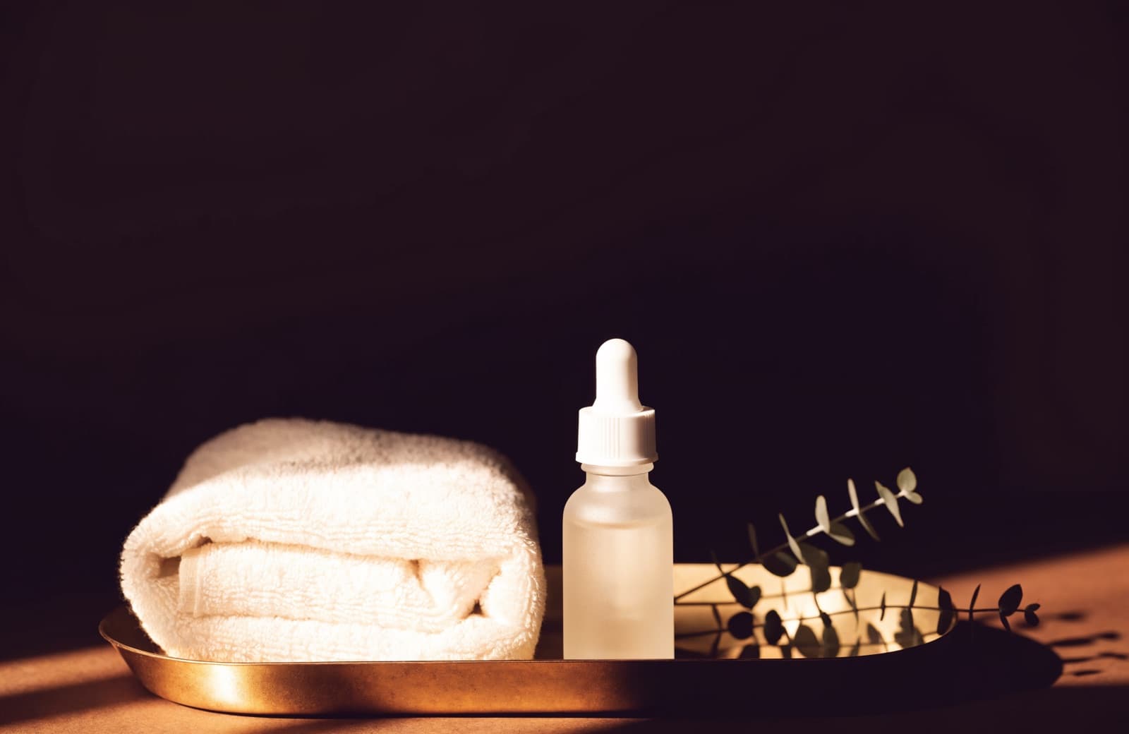 Luxury skincare still life — a folded towel, serum dropper, and eucalyptus on a gold tray